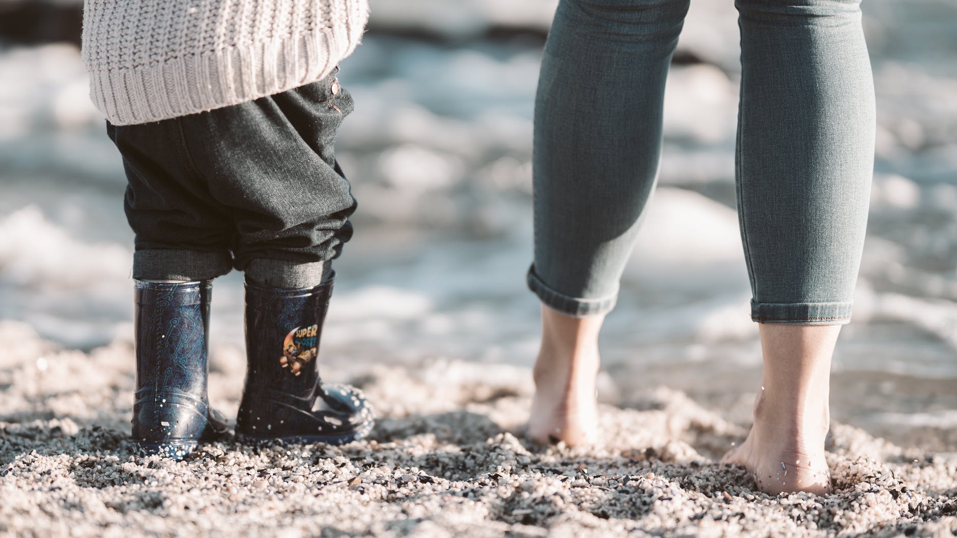 photo of adult barefoot and child on beach in wellies with thick pants on. Sea is in the background.
